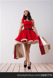 Christmas shopping. Pretty woman walking with shopping bags against white wall background, wearing red Santa Claus hat and dress