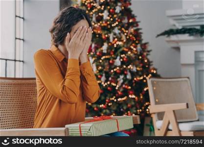 Christmas surprise concept. Curious woman covering eyes with hands and guessing what inside of gift box, sitting near New Year tree at home during winter holidays, young female getting xmas present. Curious woman covering eyes with hands and guessing what inside of Christmas gift box