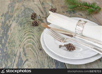 Christmas table: silver knife, fork and linen napkin lie on the white dinner plate, as well as green spruce branch and cones which is located on a old wooden table, with space for text