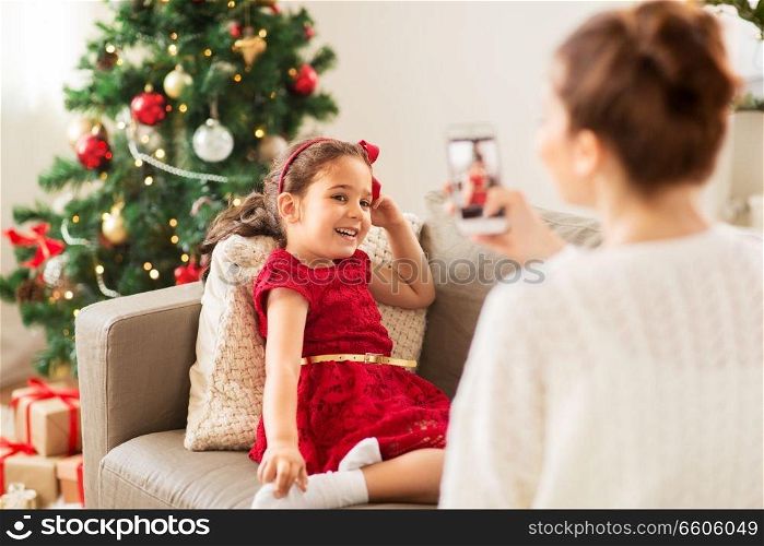 christmas, technology and family concept - mother photographing little daughter by smartphone at home. mother picturing daughter by phone on christmas