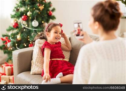 christmas, technology and family concept - mother photographing little daughter by smartphone at home. mother picturing daughter by phone on christmas