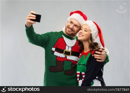 christmas, technology and holidays concept - happy couple in santa hats taking selfie by smartphone at ugly sweater party. happy couple in christmas sweaters taking selfie