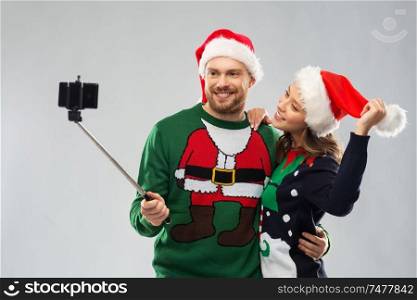 christmas, technology and holidays concept - happy couple in santa hats taking picture by smartphone on selfie stick at ugly sweater party. happy couple in christmas sweaters taking selfie