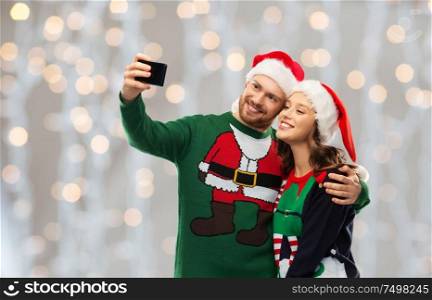 christmas, technology and holidays concept - happy couple in santa hats taking selfie by smartphone at ugly sweater party over festive lights background. happy couple in christmas sweaters taking selfie