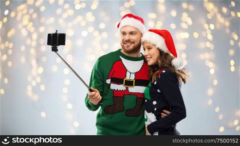 christmas, technology and holidays concept - happy couple in santa hats taking picture by smartphone on selfie stick at ugly sweater party over festive lights background. happy couple in christmas sweaters taking selfie