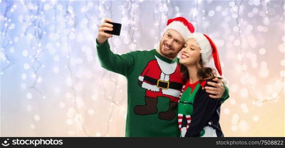 christmas, technology and holidays concept - happy couple in santa hats taking selfie by smartphone at ugly sweater party over festive lights background. happy couple in christmas sweaters taking selfie
