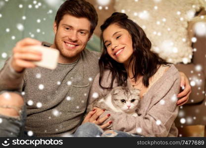 christmas, technology and people concept - happy couple with cat taking selfie by smartphone at home over snow. happy couple taking selfie by smartphone at home