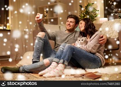christmas, technology and people concept - happy couple with cat taking selfie by smartphone at home over snow. happy couple taking selfie by smartphone at home