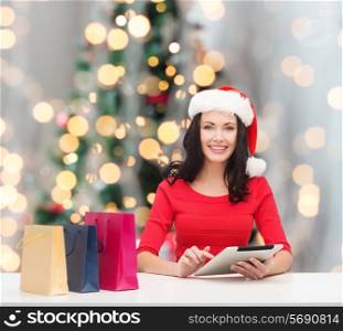 christmas, technology and people concept - smiling woman in santa helper hat with shopping bags and tablet pc computer over tree lights background