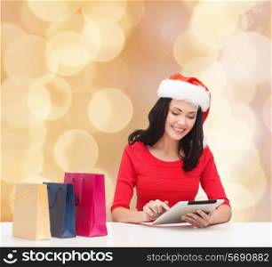 christmas, technology and people concept - smiling woman in santa helper hat with shopping bags and tablet pc computer over beige lights background