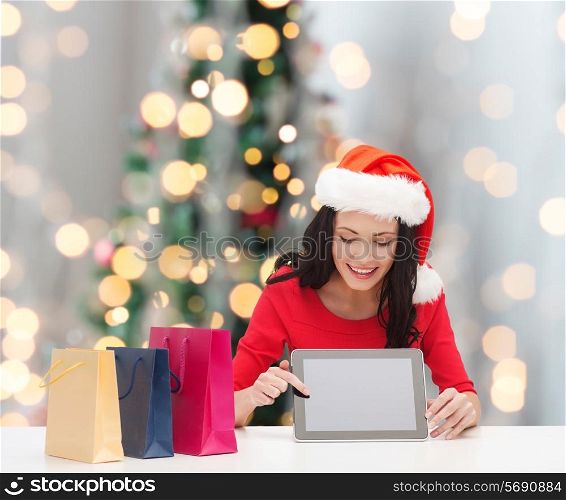 christmas, technology and people concept - smiling woman in santa helper hat with shopping bags and tablet pc computer over tree lights background