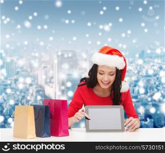 christmas, technology and people concept - smiling woman in santa helper hat with shopping bags and tablet pc computer over snowy city background