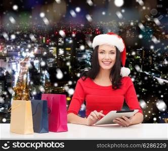 christmas, technology and people concept - smiling woman in santa helper hat with shopping bags and tablet pc computer over snowy night city background