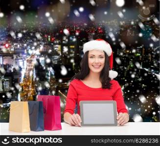 christmas, technology and people concept - smiling woman in santa helper hat with shopping bags and tablet pc computer over snowy night city background