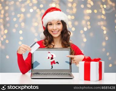 christmas, technology, people and online shopping concept - woman in santa helper hat with gift box, laptop computer and credit card over holidays lights background