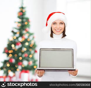 christmas, technology, winter holidays and people concept - smiling woman in santa helper hat with blank screen laptop computer over living room and christmas tree background