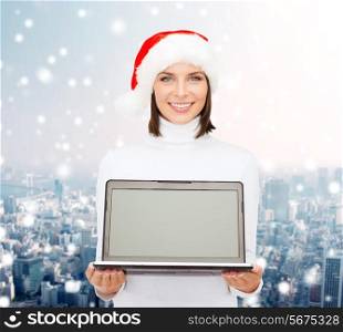 christmas, technology, winter holidays and people concept - smiling woman in santa helper hat with blank screen laptop computer over snowy city background