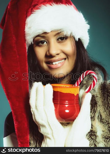 Christmas time concept. Closeup mixed race teen girl wearing santa helper hat holding red mug with hot beverage and striped candy cane studio shot on blue