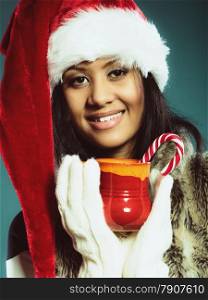 Christmas time concept. Closeup mixed race teen girl wearing santa helper hat holding red mug with hot beverage and striped candy cane studio shot on blue