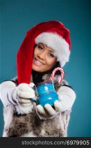 Christmas time concept. Mixed race teen girl wearing santa helper hat holding blue mug with hot beverage and striped candy cane studio shot