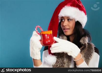 Christmas time concept. Mixed race teen girl wearing santa helper hat holding red mug with hot beverage and striped candy cane studio shot on blue