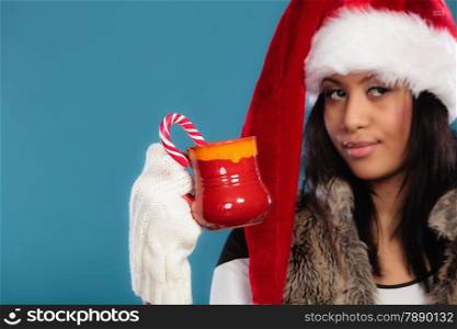 Christmas time concept. Mixed race teen girl wearing santa helper hat holding red mug with hot beverage and striped candy cane studio shot on blue
