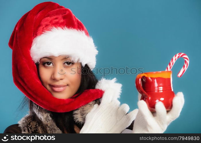 Christmas time concept. Mixed race teen girl wearing santa helper hat holding red mug with hot beverage and striped candy cane studio shot on blue