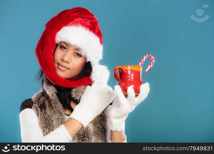 Christmas time concept. Mixed race teen girl wearing santa helper hat holding red mug with hot beverage and striped candy cane studio shot on blue