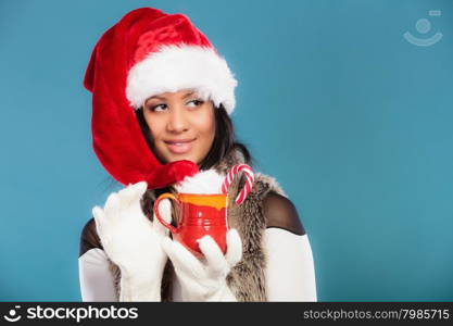 Christmas time concept. Mixed race teen girl wearing santa helper hat holding red mug with hot beverage and striped candy cane studio shot on blue