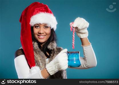 Christmas time concept. Mixed race teen girl wearing santa helper hat holding blue mug with hot beverage and striped candy cane studio shot