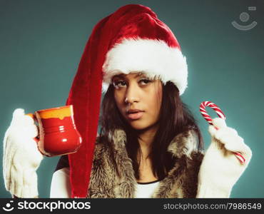 Christmas time concept. Mixed race teen girl wearing santa helper hat holding red mug with hot beverage and striped candy cane studio shot on blue