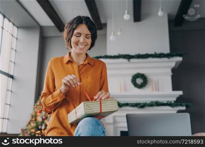 Christmas time. Happy young Spanish woman engaged in packing Xmas gifts, orders for customers, friends, enjoying winter holidays at home. Smiling european lady sitting in room decorated for New Year. Happy young Spanish woman engaged in packing Xmas gifts at home during Christmas holidays