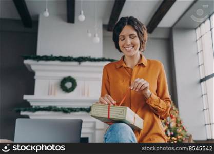 Christmas time. Happy young Spanish woman engaged in packing Xmas gifts, orders for customers, friends, enjoying winter holidays at home. Smiling european lady sitting in room decorated for New Year. Happy young Spanish woman engaged in packing Xmas gifts at home during Christmas holidays