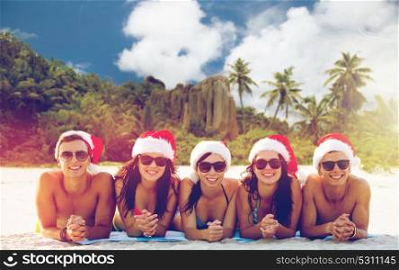 christmas, travel, winter holidays and people concept - group of friends in santa helper hats sitting over tropical seychelles island beach background. friends in santa hats on beach at christmas