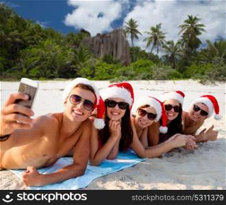 christmas, travel, winter holidays and people concept - group of friends in santa hats taking selfie by smartphone over tropical seychelles island beach background. group of friends in santa hats with taking selfie