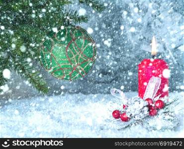 Christmas tree ball on a branch and a burning red candle under a running large snow