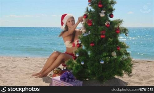 Christmas tropical beach vacations, bikini girl in Santa hat decorates a Christmas tree looking at camera