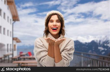 christmas, vacation and winter holidays concept - happy smiling young woman in knitted hat and woolen sweater sending air kiss at ski resort in austria over apls mountains background. woman in winter hat sending air kiss at ski resort
