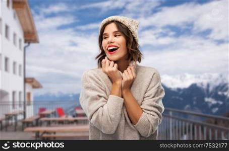 christmas, vacation and winter holidays concept - happy smiling young woman in knitted hat and woolen sweater at ski resort in austria over apls mountains background. woman in winter hat and sweater at ski resort