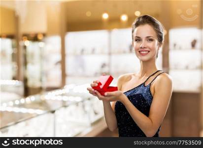 christmas, valentine’s day and luxury concept - smiling young woman in dress with gift box over jewelry store background. smiling woman with red gift box at jewelry store
