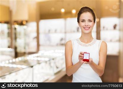 christmas, valentine’s day and luxury concept - smiling young woman in white dress with diamond ring in red gift box over jewelry store background. woman with ring in gift box at jewelry store