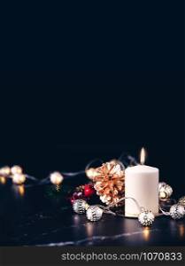 Christmas white candle with golden pine cone,mistletoe and light string glowing on black marble table with dark background.vertical banner holiday greeting card.
