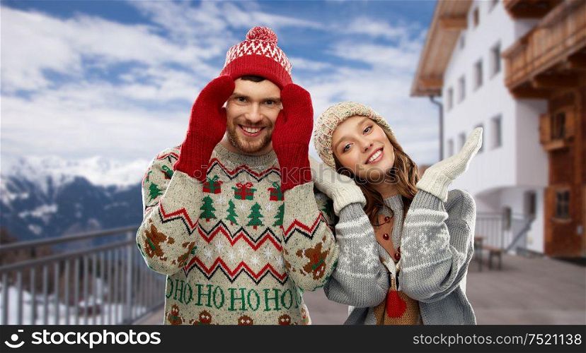 christmas, winter clothes and holidays concept - happy couple in ugly sweaters, knitted hats and mittens over ski resort in austrian alps mountains background. couple in ugly christmas sweaters over ski resort
