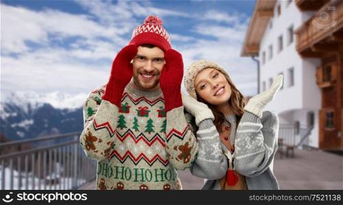 christmas, winter clothes and holidays concept - happy couple in ugly sweaters, knitted hats and mittens over ski resort in austrian alps mountains background. couple in ugly christmas sweaters over ski resort