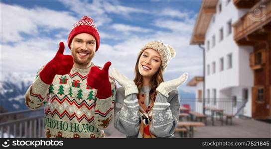 christmas, winter clothes and holidays concept - happy couple in ugly sweaters, knitted hats and mittens showing thumbs up over ski resort in austrian alps mountains background. couple in ugly christmas sweaters over ski resort