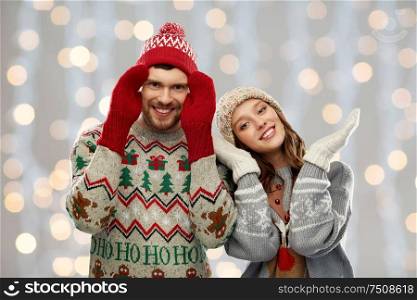 christmas, winter clothes and holidays concept - portrait of happy couple in ugly sweaters, knitted mittens and hats over festive lights background. couple in ugly sweaters and mittens on christmas