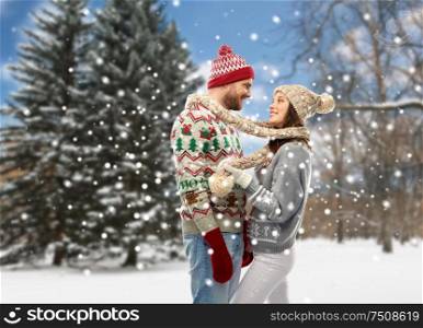 christmas, winter clothes and holidays concept - portrait of happy couple in ugly sweaters, knitted mittens and hats over snowy park background. couple in christmas ugly sweaters at winter park