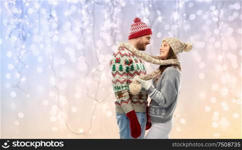 christmas, winter clothes and holidays concept - portrait of happy couple in ugly sweaters, knitted mittens and hats over festive lights background. happy couple at christmas ugly sweater party