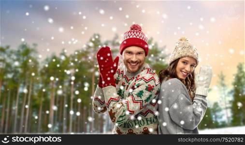 christmas, winter clothes and holidays concept - portrait of happy couple in mittens, hats and ugly sweaters over snow and forest background. couple in christmas ugly sweaters in winter