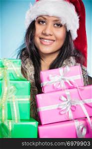 Christmas winter happiness concept. Girl mixed race woman wearing santa helper hat holding stack of presents gift boxes on blue